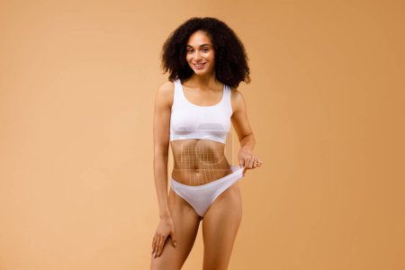 Photo for Slender african american lady wearing white underwear with net on her belly, pulling up panties, standing isolated on beige studio background. Concept of thread lifting and surgery, collage - Royalty Free Image