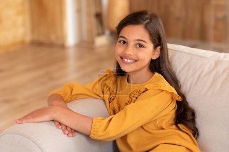 Foto de Retrato de una linda chica árabe preadolescente en una sonrisa casual mirando a la cámara, sentada en un sofá en casa. Preadolescente niño expresando emociones positivas posando en la sala de estar en interiores - Imagen libre de derechos