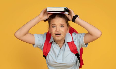 Foto de Sorprendido triste adolescente europea con mochila sostiene libro en la cabeza, sufre de exceso de trabajo y estrés en la escuela, aislado en fondo de estudio naranja. Problemas con el estudio, el conocimiento, los deberes - Imagen libre de derechos