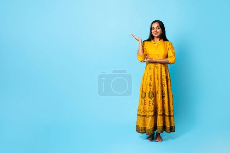 Foto de Longitud completa de la joven india feliz en vestido amarillo tradicional gestos con la mano, mostrando el espacio de copia, publicidad de moda o la oferta de belleza de pie sobre fondo de estudio azul - Imagen libre de derechos