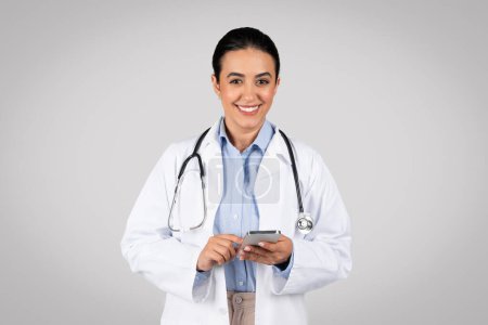 Photo for Portrait of friendly latin female therapist in white coat and with stethoscope using cellphone for work and smiling at camera, gray background. Health care and treatment remotely, app for medicine - Royalty Free Image