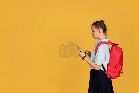 Foto de Adolescente europea seria colegiala con mochila escribiendo en el teléfono en la escuela, aislado en fondo de estudio naranja, perfil. Redes sociales, aplicación para el estudio, conocimiento, lección en línea - Imagen libre de derechos