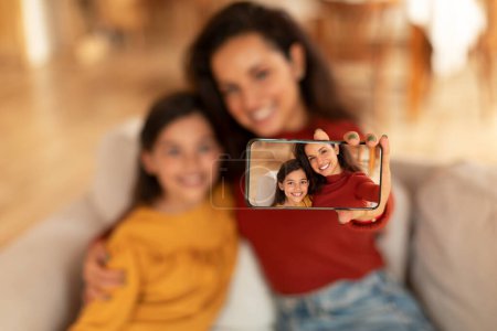 Foto de Happy Middle Eastern Young Mom and Daughter Making Selfie Photo Via Phone, Abrazándose en el sofá en casa, Enfoque selectivo en la pantalla del teléfono inteligente con disparo de madre feliz y niña. Profundidad superficial - Imagen libre de derechos