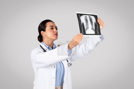 Téléchargez les photos : Jeune médecin latino-américaine tenant et regardant la radiographie pulmonaire, debout sur fond de studio gris avec une expression confiante, pensant sérieusement - en image libre de droit