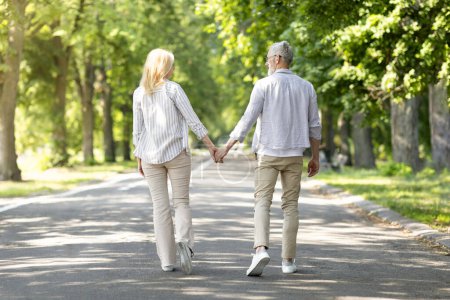 Photo for Outdoor Date. Rear View Of Romantic Mature Couple Walking Together In Park, Happy Loving Senior Man And Woman Holding Hands And Smiling To Each Other, Enjoying Time With Each Other, Copy Space - Royalty Free Image
