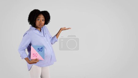 Photo for Confused young black lady with belly hold banner with 50 percent boy or girl sign and free space on hand, isolated on gray background, studio. Gender party, ad and offer, question - Royalty Free Image