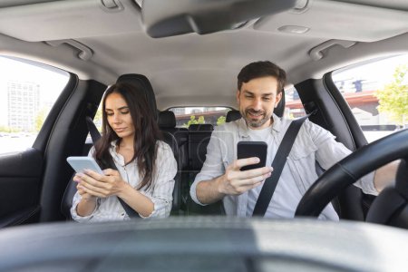 Photo for Millennial man and woman sitting inside car on front seats, using smartphones. Couple going somewhere by auto, checking their cell phones, dont talk to each other. Unsafe driving concept - Royalty Free Image