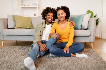 Téléchargez les photos : Épouses afro-américaines romantiques étreignant assis sur le sol près du canapé à la maison, exprimant des émotions positives souriant à la caméra dans le salon intérieur moderne. Mari embrasse sa jeune femme - en image libre de droit