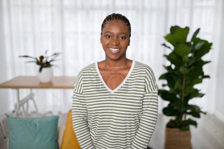 Foto de Retrato de alegre joven afroamericana feliz con traje casual sentado en el sofá en la acogedora sala de estar soleada, sonriendo a la cámara, dama negra posando sola en casa, espacio para copiar - Imagen libre de derechos