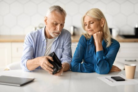 Photo for Financial Crisis. Upset Senior Spouses Looking At Empty Wallet While Sitting At Table In Kitchen, Mature Husband And Wife Suffering Retirement Problems, Having Economy Issues And Money Absence - Royalty Free Image