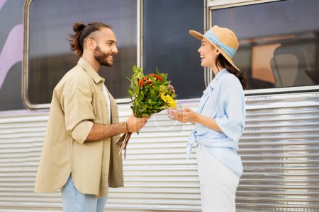 Photo for Cheerful young caucasian guy meets surprised woman, gives bouquet of flowers on train station. Hello, love, relationship, travel and lifestyle, dating and romance, vacation - Royalty Free Image