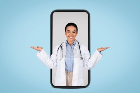 Photo for Cheerful young brazilian woman doctor holding something invisible on both hands palms, posing in huge cellphone screen isolated on blue studio background - Royalty Free Image