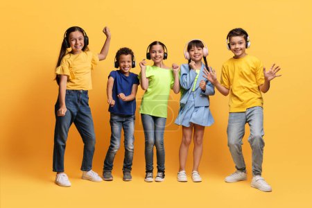 Funny multiracial diverse kids listening to music and dancing, using wireless headphones. Happy children preteen boys and girls having fun isolated on yellow background. Kids entertainment