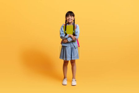Téléchargez les photos : Jolie pré-adolescente chinoise portant un uniforme bleu allant à l'école, portant un sac à dos, tenant des cahiers d'exercices, isolée sur fond jaune, espace de copie. éducation des enfants, école - en image libre de droit