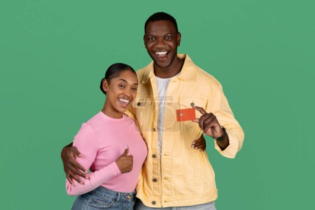 Foto de Emocionado feliz millennial negro hombre y mujer sosteniendo tarjeta de crédito de plástico rojo con chip, sonriendo y mostrando el pulgar hacia arriba, aislado sobre fondo verde. Banca, depósito, préstamo, compras, venta minorista - Imagen libre de derechos