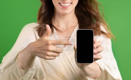 Foto de Feliz joven europea dama señalar el dedo en el teléfono inteligente con pantalla en blanco, aislado en fondo de estudio verde, de cerca. Recomendación de venta, sitio web y aplicación para compras, anuncios y ofertas - Imagen libre de derechos