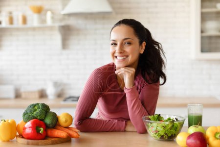 Téléchargez les photos : Portrait de jeune femme heureuse en Fitwear posant à table avec des légumes et des fruits biologiques frais dans la cuisine moderne à l'intérieur, souriant à la caméra. Healthy Weight Loss Diet Recipes Concept - en image libre de droit