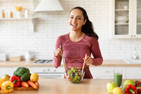 Photo for Healthy weight loss nutrition. Joyful slim lady in sportswear cooking fresh vegetable salad, smiling to camera while mixing ingredients standing at kitchen table at home interior. Slimming recipes - Royalty Free Image