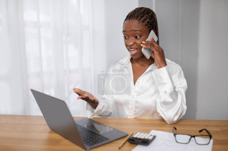 Téléchargez les photos : Jeune femme noire en colère dans les tenues formelles gestionnaire assis à la table, regardant l'écran d'ordinateur portable et des gestes, femme d'affaires ont convetsation téléphone avec assistant, intérieur de bureau, espace de copie - en image libre de droit