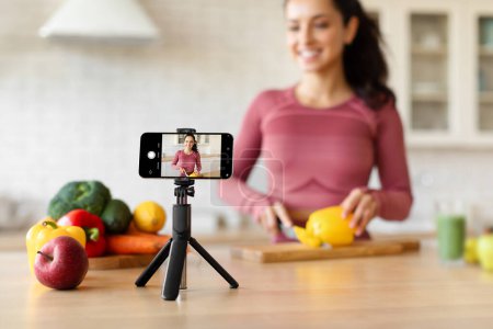 Photo for Healthy Nutrition Blogging. Smartphone With Fitness Blogger On Screen, Shooting Video For Social Media While Woman Cooking Healthy Meal For Weight Loss At Kitchen. Shallow Depth, Focus On Phone - Royalty Free Image