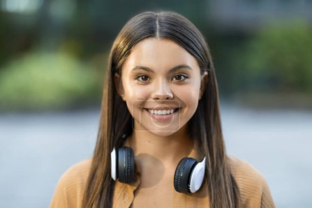 Photo for Portrait of cheerful pretty long-haired young lady student posing outdoors at university campus, wearing casual outfit and wireless headphones, smiling at camera. Generation z, zoomers lifestyle - Royalty Free Image