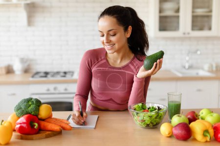 Photo for Weight Loss Diet Plan. Smiling Fitness Woman Writing Down Healthy Recipes In Her Notebook, Holding Avocado, Planning Daily Meals At Kitchen Table With Fresh Vegetables And Fruits Indoor. Nutrition - Royalty Free Image