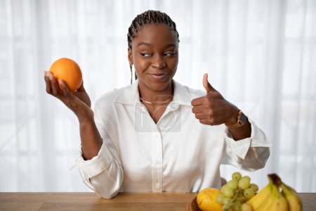 Foto de Positiva feliz atractiva joven mujer negra en camisa blanca dietista con naranja jugosa fresca en la mano, mostrando el pulgar hacia arriba y sonriendo, recomendando comida saludable llena de vitaminas, espacio para copiar - Imagen libre de derechos