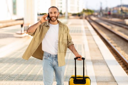Photo for Positive millennial caucasian guy with suitcase calling by phone, enjoy trip at train station. Active lifestyle, summer vacation and homecoming, business travel, communication - Royalty Free Image