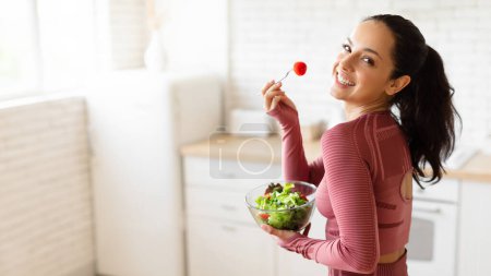 Téléchargez les photos : Facile et savoureux minceur. Femme brune joyeuse en vêtements de sport posant manger salade végétarienne, tenant bol et profiter d'un dîner sain à l'intérieur de la cuisine moderne. Panorama avec espace libre pour le texte - en image libre de droit