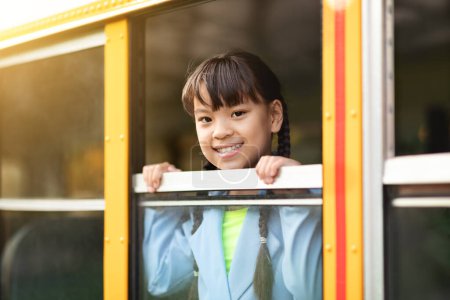 Photo for Cute preteen asian girl looking out of school bus window, cheerful female child smiling at camera while peeking out of yellow schoolbus, ready for trip to home after lessons, closeup shot - Royalty Free Image