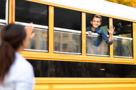 Foto de Madre saludando de la mano al hijo sentado en el autobús escolar amarillo, niño preadolescente feliz diciendo adiós a mamá por la mañana mientras va a clases en autobús escolar, alumno masculino disfrutando de viaje a la escuela, enfoque selectivo - Imagen libre de derechos