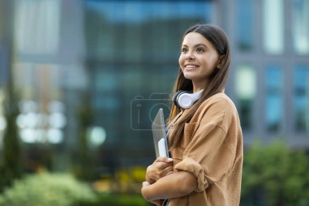 Foto de Feliz guapa morena de pelo largo joven estudiante posando en la universidad o campus universitario al aire libre, llevando portátil y teléfono inteligente, mirando el espacio de copia y sonriendo. Concepto de educación en línea - Imagen libre de derechos