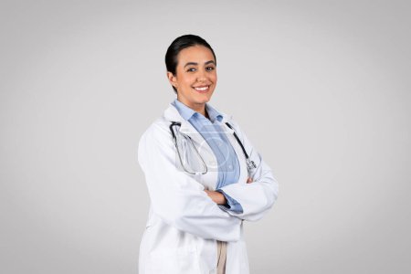 Téléchargez les photos : Travailleur médical. Joyeux médecin brésilien femme en uniforme blanc souriant à la caméra tout en posant sur fond gris, thérapeute féminine avec stéthoscope - en image libre de droit