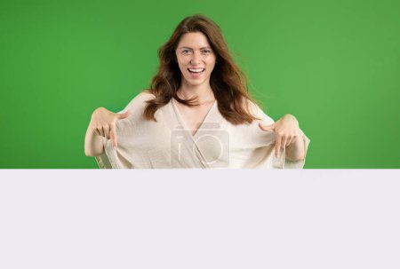 Photo for Smiling pretty young european lady in casual pointing fingers at large banner with empty space, isolated on green studio background. Sale, recommendation for work and study, ad and offer - Royalty Free Image