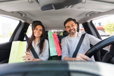 Photo for Happy beautiful millennial couple driving home after shopping in mall, smiling. Cheerful man and woman sitting inside car, holding colorful shopping bags purchases. Black friday, season sale - Royalty Free Image
