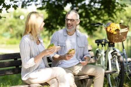 Téléchargez les photos : Heureux couple de personnes âgées mangeant des sandwichs tout en étant assis sur le banc dans le parc, Romantique Mature conjoints déjeunant à l'extérieur et riant ensemble, joyeux homme et femme plus âgés appréciant pique-nique à l'extérieur - en image libre de droit