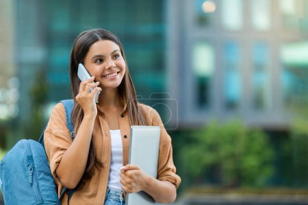 Photo for Cheerful happy pretty young woman student carrying backpack and laptop in her hand have phone call with friend, walking by university campus, looking at copy space and smiling. Students communication - Royalty Free Image