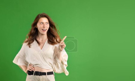 Téléchargez les photos : Heureux surpris jeune femme européenne décontractée à bouche ouverte, pointant du doigt l'espace de copie, isolé sur fond de studio vert. Choix, question et idée, émotions humaines, publicité et offre - en image libre de droit