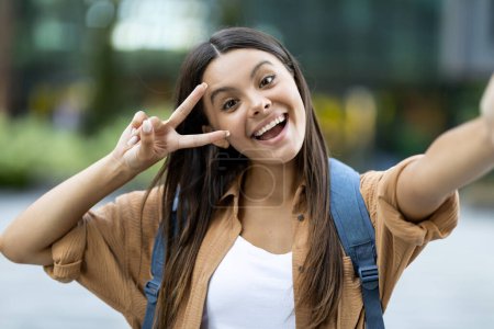 Foto de Linda morena de pelo bastante largo joven zoomer tomar selfie al aire libre, haciendo gestos de paz y sonriendo, caminando por la calle, ir a la universidad, universidad, publicar en las redes sociales, espacio para copiar - Imagen libre de derechos
