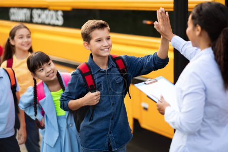 Téléchargez les photos : Enseignante noire donnant High Five au garçon entrant dans le bus scolaire, assistante femme interagissant avec les enfants avant le voyage, contrôlant la liste de contrôle des élèves entrant dans Shoolbus, foyer sélectif - en image libre de droit