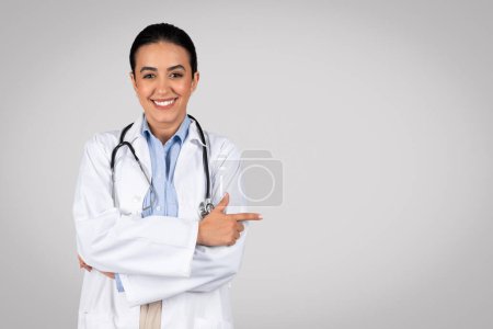 Foto de ¡Míralo! Feliz doctora latina en el espacio de publicidad de ropa de trabajo blanca para la publicidad, señalando el dedo a un lado en el espacio de copia, de pie sobre el fondo gris del estudio, espacio para la oferta médica - Imagen libre de derechos