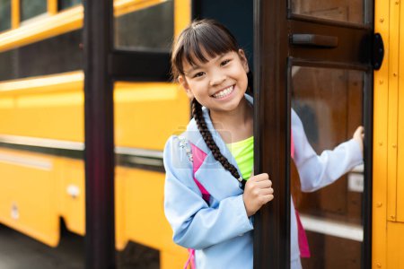 Foto de Linda chica asiática de pie en la puerta del autobús escolar y mirando a la cámara, niño preadolescente alegre asomándose desde el autobús escolar amarillo, listo para el viaje a casa después de las lecciones, primer plano con espacio de copia - Imagen libre de derechos