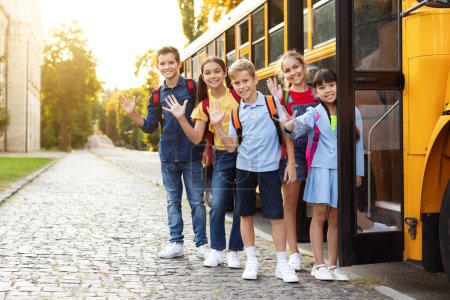 Photo for Happy Multiethnic Kids Waving Hand At Camera While Boarding School Bus, Cheerful Schoolboys And Schoolgirls With Backpacks Having Trip Together, Going Home After Classes, Full Length, Copy Space - Royalty Free Image