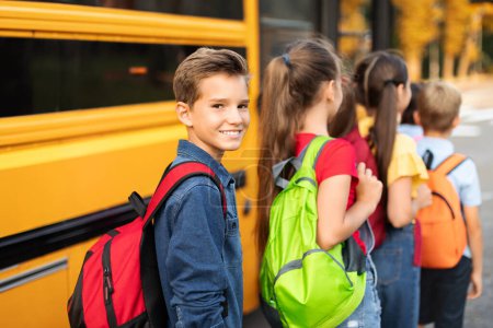 Foto de Retrato de niño sonriente con mochila abordaje autobús escolar amarillo con compañeros de clase y mirando a la cámara, Grupo de niños que entran en el vehículo después de lecciones, listos para el viaje juntos, Primer plano - Imagen libre de derechos
