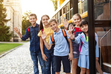Foto de Niños multiétnicos felices parados cerca del autobús escolar y saludando con la mano en la cámara, alumnos alegres con mochilas listos para el viaje a casa después de lecciones, niños alegres disfrutando del transporte escolar - Imagen libre de derechos