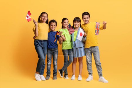Foto de Los niños sonrientes con banderas de diferentes países en las manos disfrutan de la educación internacional. Feliz escuela multiétnica niños en edad de participar en la experiencia multirracial, fondo amarillo - Imagen libre de derechos