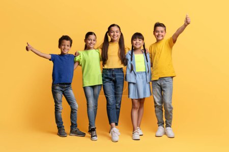 Photo for Cool cheerful diverse multiracial preteen kids having fun isolated on yellow background, embracing, showing thumb up and smiling at camera. School aged boys and girls posing together, full length - Royalty Free Image