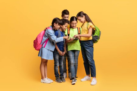 Photo for Cute multiethnic children happy boys and girls with backpacks holding digital tablet isolated on yellow background. Excited diverse kids checking nice educational website, application - Royalty Free Image