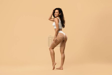 Photo for Young slim woman posing in white underwear over beige studio background, beautiful female showing perfect slender body and buttocks without cellulite, attractive lady smiling at camera, full length - Royalty Free Image