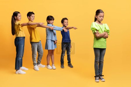 Photo for Bullying at school age concept. Diverse laughing school aged kids pointing at upset girl, isolated on yellow background. Poor child suffering from mockery from her classmates - Royalty Free Image